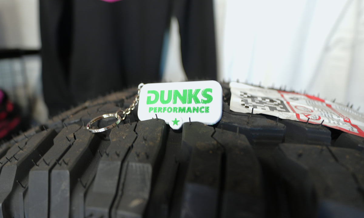 A white rectangular keychain tag with green 'DUNKS PERFORMANCE' text and a silver key ring, resting on the tread of a black off-road tire.