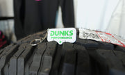 A white rectangular keychain tag with green 'DUNKS PERFORMANCE' text and a silver key ring, resting on the tread of a black off-road tire.