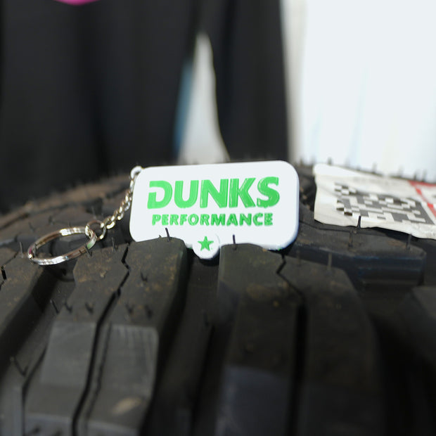A white rectangular keychain tag with green 'DUNKS PERFORMANCE' text and a silver key ring, resting on the tread of a black off-road tire.