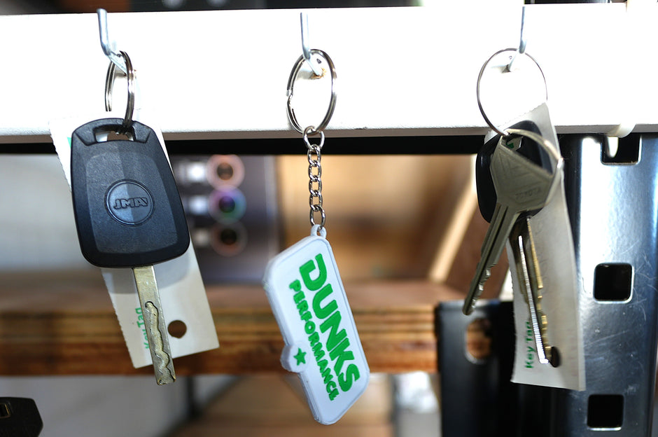 A white plastic keychain with green 'DUNKS PERFORMANCE' text hangs from a metal rack alongside other keys, including a black car key and metal keys.